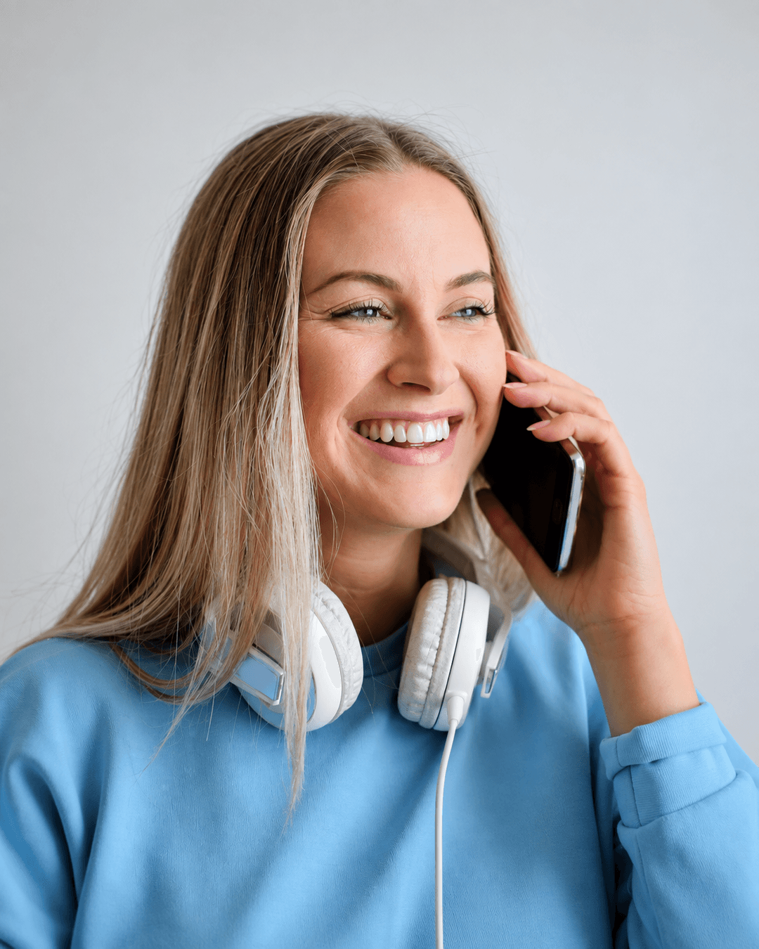 Eine lächelnde Frau im blauen Oberteil telefoniert mit dem Smartphone und trägt Kopfhörer um den Hals.